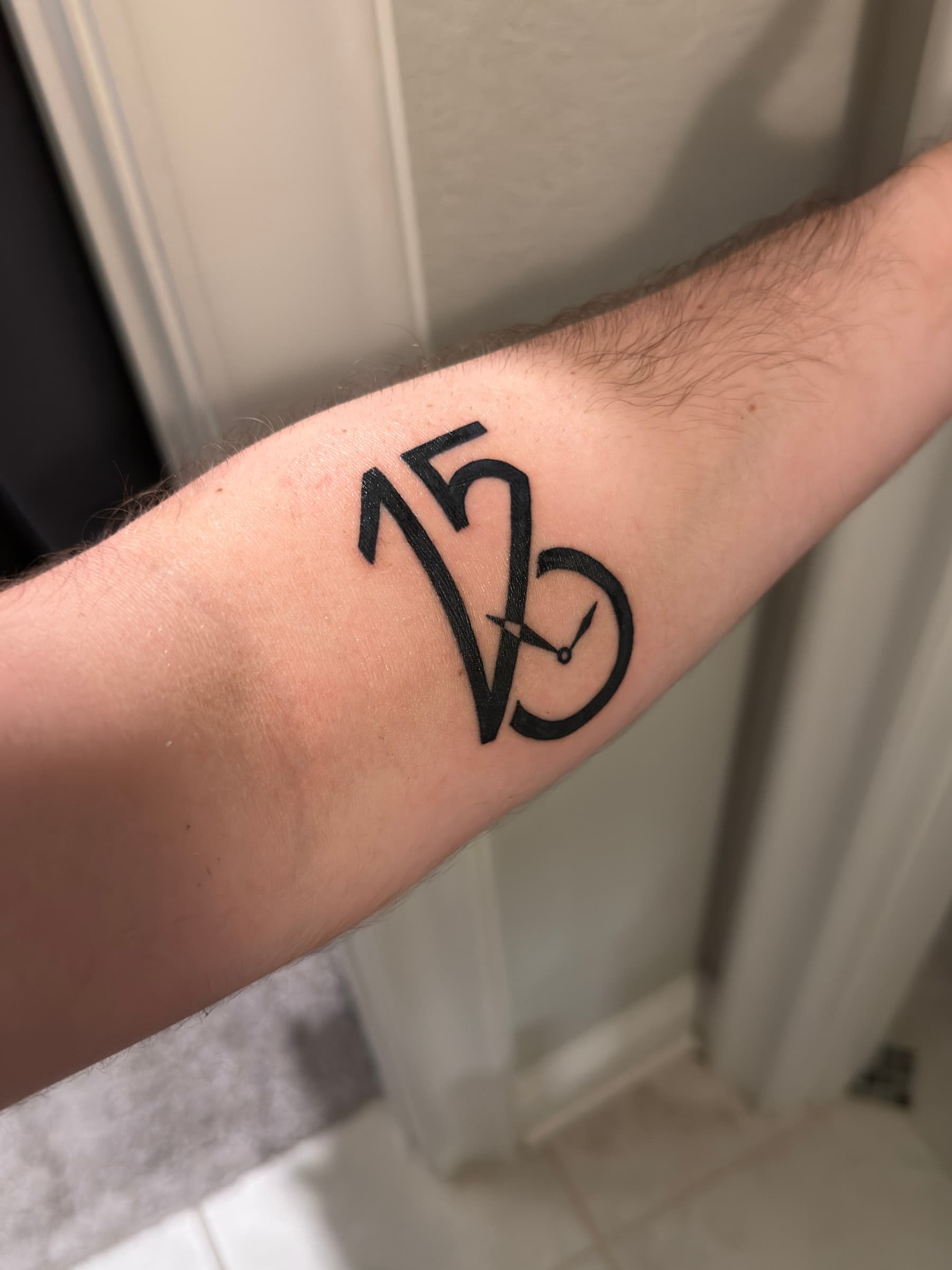 Finished black-line tattoo on the inner forearm, showing the interlocked 1·5·6, V, and watch form.